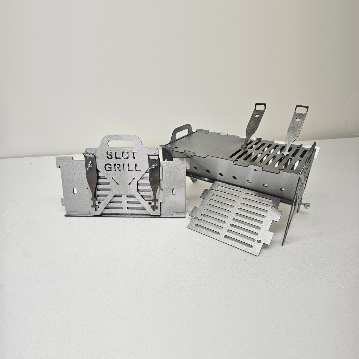 Slot Grill Compact assembled with a griddle plate and grate, next to its flat-packed panels